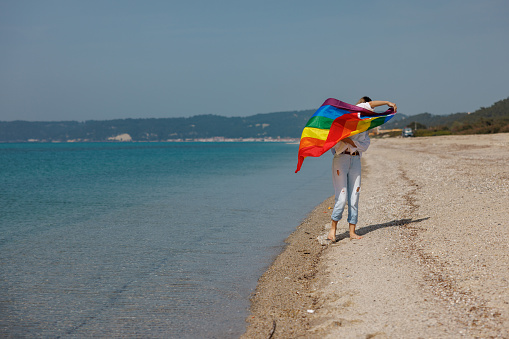 Full length shot of beautiful lesbian woman holding LGBTQ flag at the beach, concept of love and diversity