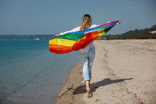 Rear view shot of young woman holding LGBTQ flag and running at the beach, concept of tolerance and diversity