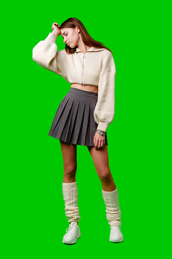 A young woman stands with one hand raised to her forehead, wearing a casual white sweater paired with a grey pleated skirt. Her outfit is completed with white socks and sneakers, conveying a relaxed yet fashionable look. The green background adds a striking contrast to the scene.