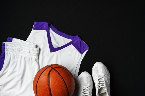 A white and purple basketball jersey and matching shorts are neatly placed beside a basketball and a pair of white sneakers, all set against a dark backdrop. The arrangement suggests preparation for a game or practice session.