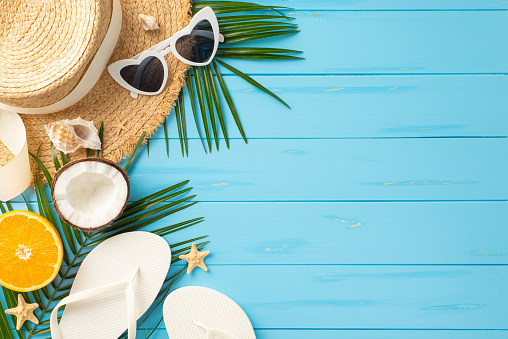 A vibrant collection of summer beach items like sunglasses, hat, flip flops, coconut, and orange on a turquoise wooden surface depicts the ideal vacation vibe