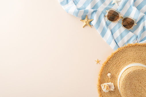Top-down view of summer relaxation scene. Sun hat, shades, beach blanket, seashells on sandy beige. Suitable for promotions or captions