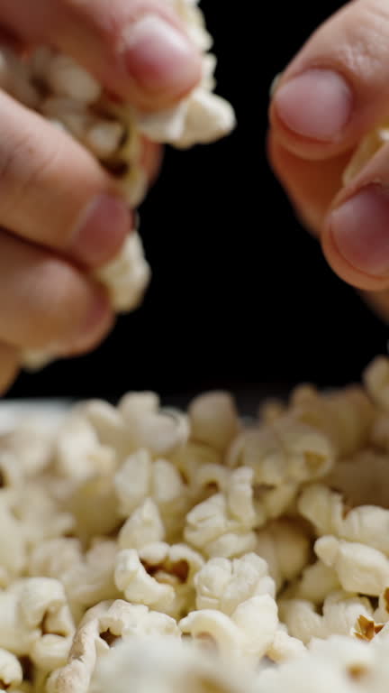 Vertical video. Hands appear from darkness on both sides and take handfuls of popcorn. Close-up, slow motion.