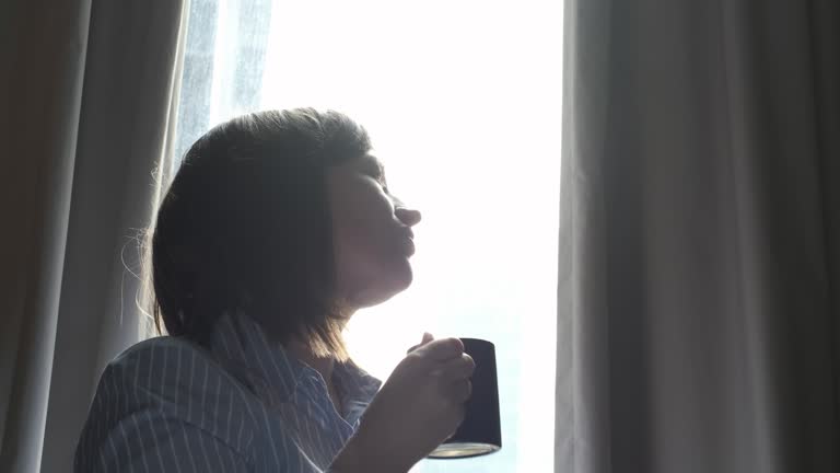 Young woman looking out window and drinking coffee side view.