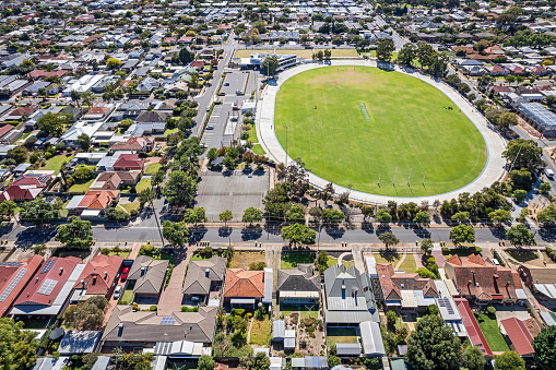 Aerial view over established houses in Adelaide suburb with nearby sports field: the grassed field (oval) has cycling track (velodrome), cricket pitch, Australian Rules Football goal posts, modern clubrooms, pavilion & off street parking.