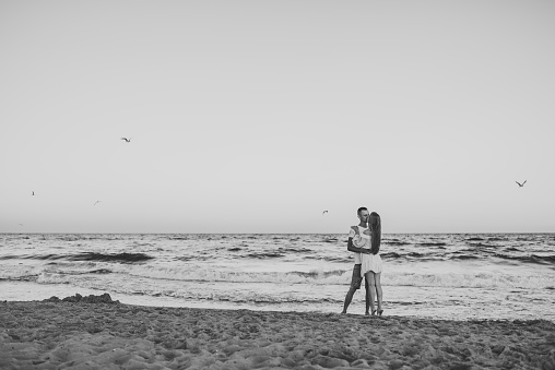Man embraces woman walks on sand sea. Couple in love hugging and kissing on seashore. Female kisses male standing barefoot on beach with big waves ocean and enjoying summer day. Black and white photo