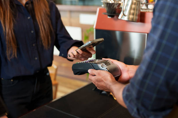 istock Customer using smartphone to complete digital payment with barista's handheld device in a cafe. Modern payment being made via mobile phone by woman in casual clothes to attentive worker in coffee shop 2150257560