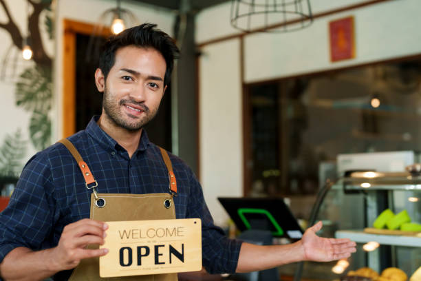 istock Young Asian man in smart cafe uniform with 'Open' sign, customer interaction, warmly lit eatery with pastries. Attentive shop assistant presenting welcoming sign, interacting with visitors, 2150257565