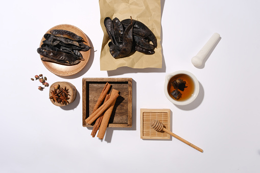 A group of raw materials evenly spread on white table, a rectangle wooden tray contains cinnamon, some locust fruit placed on brown paper and a round dish, a wooden podium with anise on. Template for designing or advertising