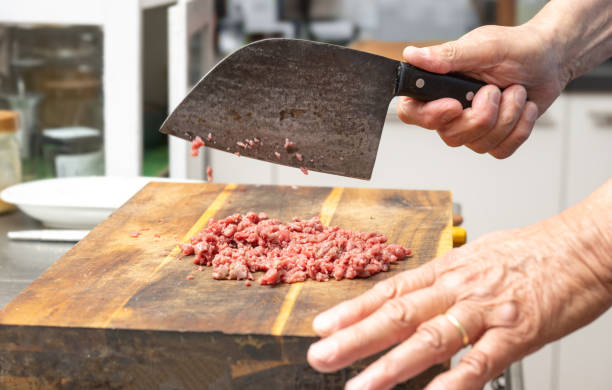 Cook chopping raw beef with a rustic kitchen cleaver on a wooden butcher block to make tartare, ground meat or minced meat, copy space, selected focus