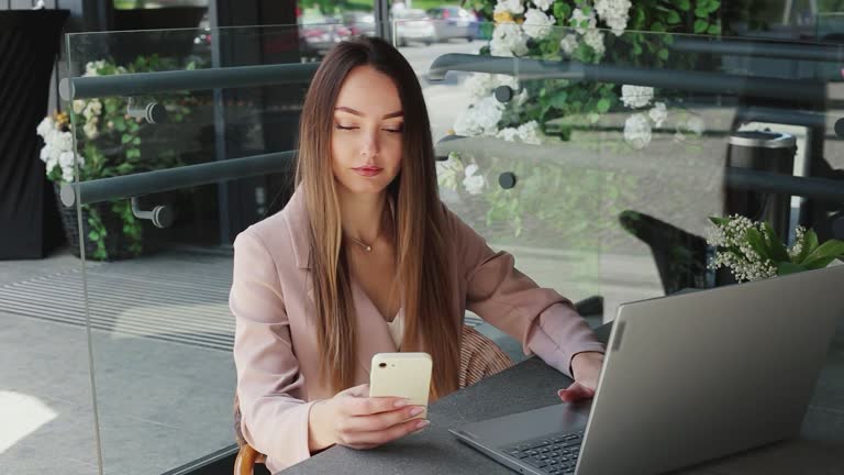 Young caucasian woman in a business suit sits and works in a cafe with a phone and a laptop