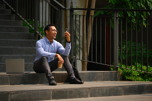 Cheerful male office worker sitting on stairs with his laptop in the city and using mobile phone.
