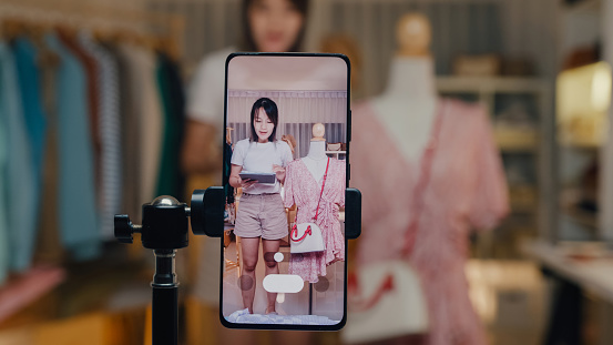 Closeup of young Asian woman live streaming on phone show new collection handbag to customer in shop at night. SME small business e-commerce concept.