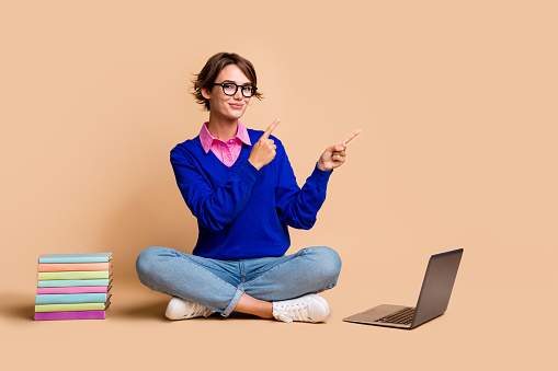 Full body portrait of smart lady sit floor book laptop indicate fingers empty space isolated on beige color background.