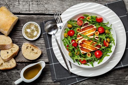 healthy salad with grilled halloumi greek cheese, arugula, tomatoes and green olives in white bowl on rustic wooden table with bread and olive oil, horizontal view from above, flat lay