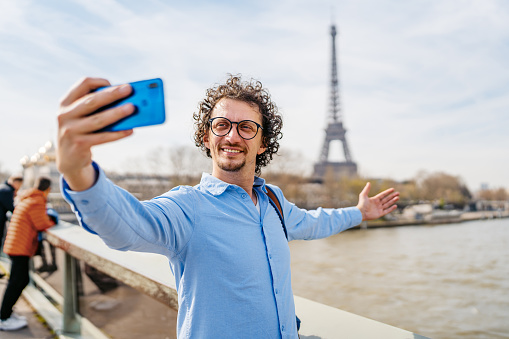 Handsome young man taking selfies using his smart phone on the bridge in Paris in France. View of the Eiffel Tower in the distance.