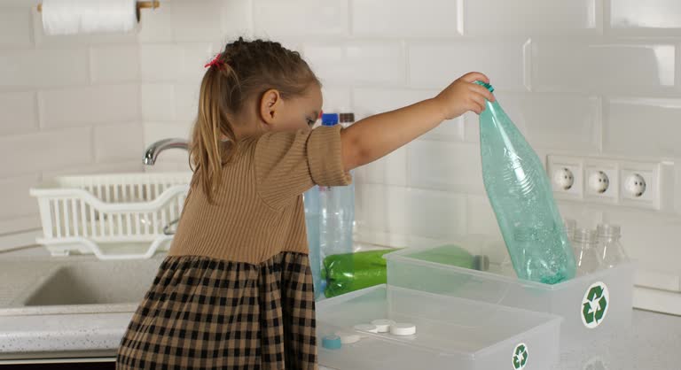 In the kitchen, a child girl puts plastic waste in a box with a recycling sign. A child takes part in his family's plastic sorting. Cute child having fun sorting out bottles for recycling.