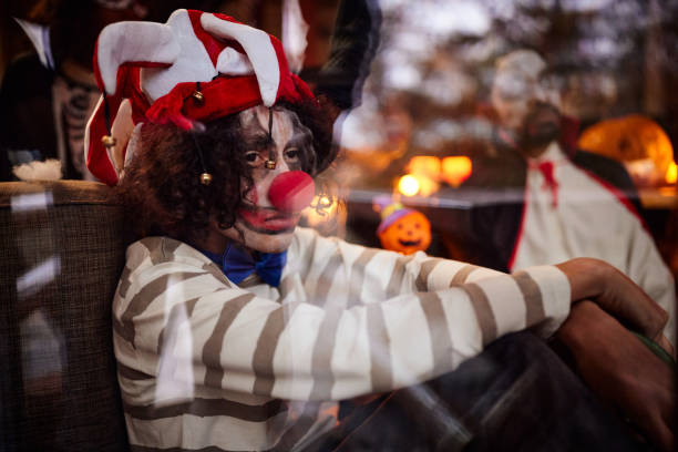 Displeased African American boy in clown costume on Halloween at home. The view is through glass. There are people in the background.