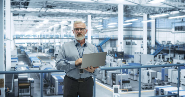 istock Portrait of a Handsome Middle Aged Engineer with a Beard and Glasses Using Laptop Computer and Looking at Camera. Male Specialist Working in a Modern Electronics Factory 2150271790
