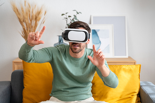 Enthusiastic young adult male navigating imaginary interface with virtual reality headset, seated on couch in modern living room setting