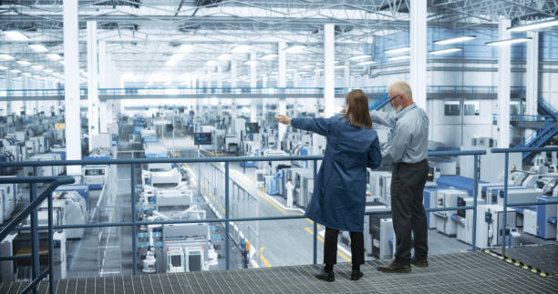 experienced male and female engineers standing on a platform with their back to camera, using laptop computer and discussing production at a modern ai automated electronics manufacture - fábrica imagens e fotografias de stock