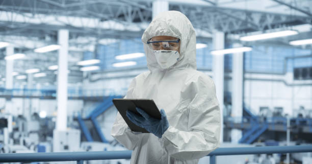 istock Female Technician in Protective Coveralls, Mask and Eyewear Using Tablet Computer at a Manufacturing Complex. Specialist Monitoring Conditions at a Modern Electronics Factory with Automated Robots 2150276083
