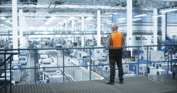 istock Industrial Engineer Working at a Technology Factory. Middle Aged Male Using Laptop Computer at Work, Browsing Electronics Information Online. Man's Back in Safety Vest Turned to Camera 2150276108