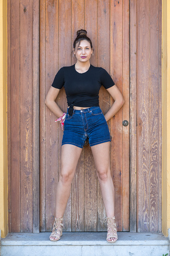 Pretty young woman in casual clothes posing with her arms behind her back with a brown door in the background