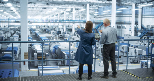 istock Male and Female Engineers Standing with Their Back to Camera, Using Laptop Computer and Talking in a Factory Facility with Equipment Producing Modern Electronic Components for Different Industries 2150276205