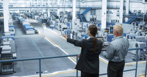 istock Male Engineer and Female Project Manager Standing on a Platform, Talking, Using Laptop Computer and Monitoring Production at a Modern Automated Electronics Manufacture with AI Robotic Arms 2150276215