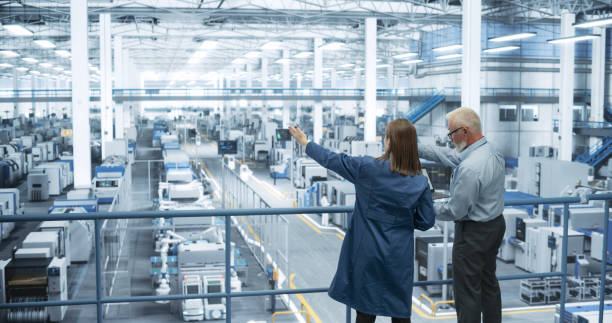 istock Male Engineer and Female Project Manager Standing on a Platform, Talking, Using Laptop Computer and Monitoring Production at a Modern Automated Electronics Manufacture with AI Robotic Arms 2150276235