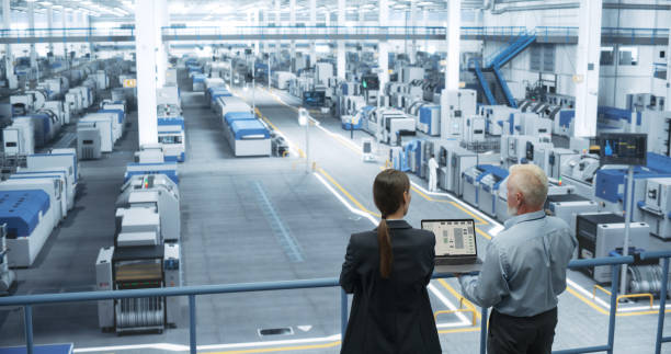 istock Male Engineer and Female Project Manager Standing on a Platform, Talking, Using Laptop Computer and Monitoring Production at a Modern Automated Electronics Manufacture with AI Robotic Arms 2150276266