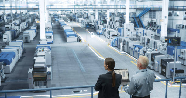 istock Middle Aged Engineer and Female Scientist Standing with Their Back, Using Laptop Computer and Talking in a Factory Facility with Equipment Producing Modern Technical Devices for Different Industries 2150276302