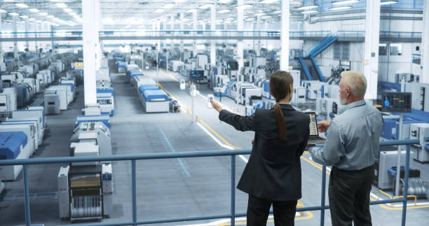 istock Middle Aged Engineer and Female Scientist Standing with Their Back, Using Laptop Computer and Talking in a Factory Facility with Equipment Producing Modern Technical Devices for Different Industries 2150276337