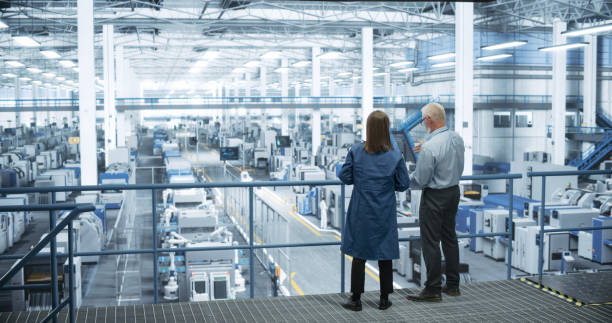 istock Middle Aged Engineer and Female Scientist Using Laptop Computer and Talking in a Factory Facility with Equipment Producing Modern Electronic Components for Different Industries 2150276360