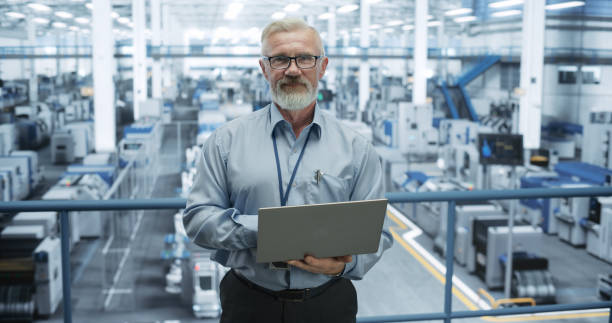 istock Portrait of a Handsome Middle Aged Engineer Wearing Glasses, Using Laptop Computer and Looking at Camera. Technician in High-VIsibility Vest Working in a Modern Technology Factory 2150276497
