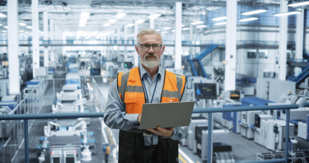 istock Portrait of a Handsome Middle Aged Engineer Wearing Glasses, Using Laptop Computer and Looking at Camera. Technician in High-VIsibility Vest Working in a Modern Technology Factory 2150276517