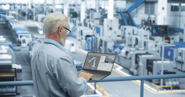 istock Portrait of a Handsome Middle Aged Engineer with Glasses Using Laptop Computer and Looking Around a Factory Facility with Equipment Producing Modern Electronic Components for Different Industries 2150276526