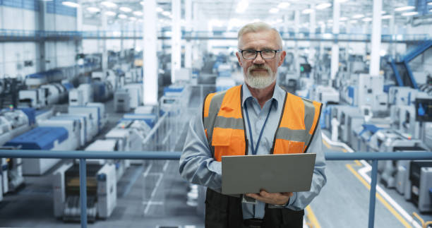 istock Portrait of a Senior Technician with a Beard and Glasses Using Laptop Computer and Looking at Camera. Industrial Male Specialist Wearing a Protective Vest, Working in a Modern Electronics Factory 2150276584