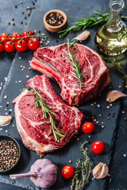 istock Two raw beef steaks on the bone on a black slate serving plate, decorated with various ingredients. 2150281980