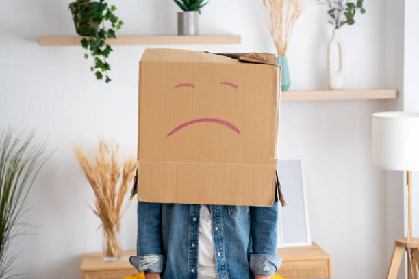 Individual stands indoors with a cardboard box on their head, featuring a hand-drawn sad face, symbolizing anonymity or emotion concealment