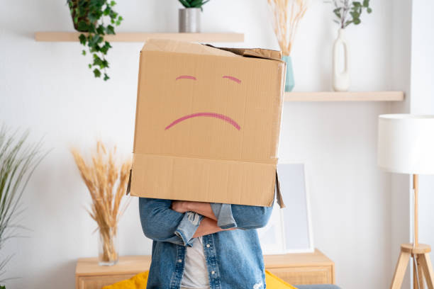 Person standing in a living room holding a cardboard box with a sad face drawn on it over their head
