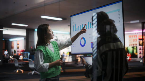 Two Creative Colleagues Collaborating, Pointing at Television Set With Financial Graphs And Charts in Office. Asian Female CEO Consults Black Marketing Manager. Two Women Brainstorming