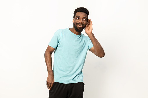 A cheerful young man in a blue t-shirt cupping his ear to listen attentively against a white background.