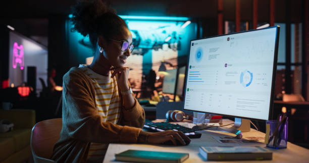 Female Talented Digital Marketing Specialist Working on Computer at Office in the Evening. Young Black Businesswoman in Creative Agency, Smiling and Researching Business Analytical Data