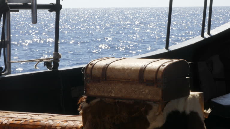 Pirate treasure chest atop a viking fur pelt inside a longboat galleon ship, with sparkling sea outside. Evokes piracy, treasure hunting, and historical sea voyages