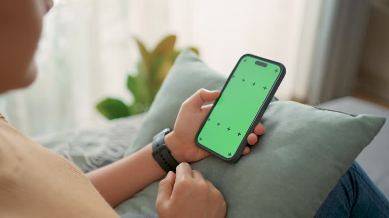 Woman holding smartphone and sliding with green screen mock-up sitting on sofa at home.
