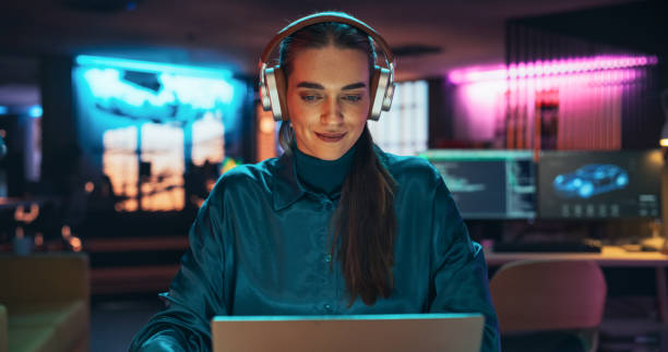 Young Project Manager with Headphones Working on Laptop Computer in Creative Office Environment. Beautiful Female Specialist is Writing Business Strategy, Using Digital Tablet to Take Notes