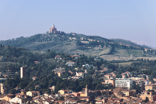 A shot from Bologna city center capturing the distant Santuario della Madonna di San Luca atop a hill. The cityscape below includes tightly grouped buildings and terracotta rooftops