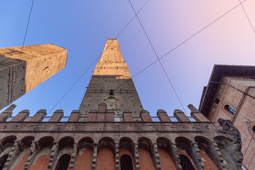 The angular perspective showcases the Asinelli and Garisenda towers in Bologna, reaching skyward, framed by architectural details and intersecting power lines against a fading sky